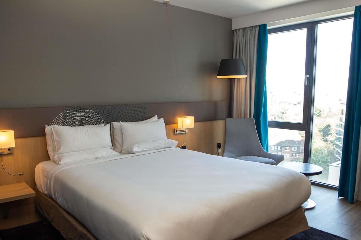 Hotellihuone, jossa on suuri sÀnky, kaksi yöpöytÀÀ, tuoli, ikkuna verhoilla ja lamppu.
