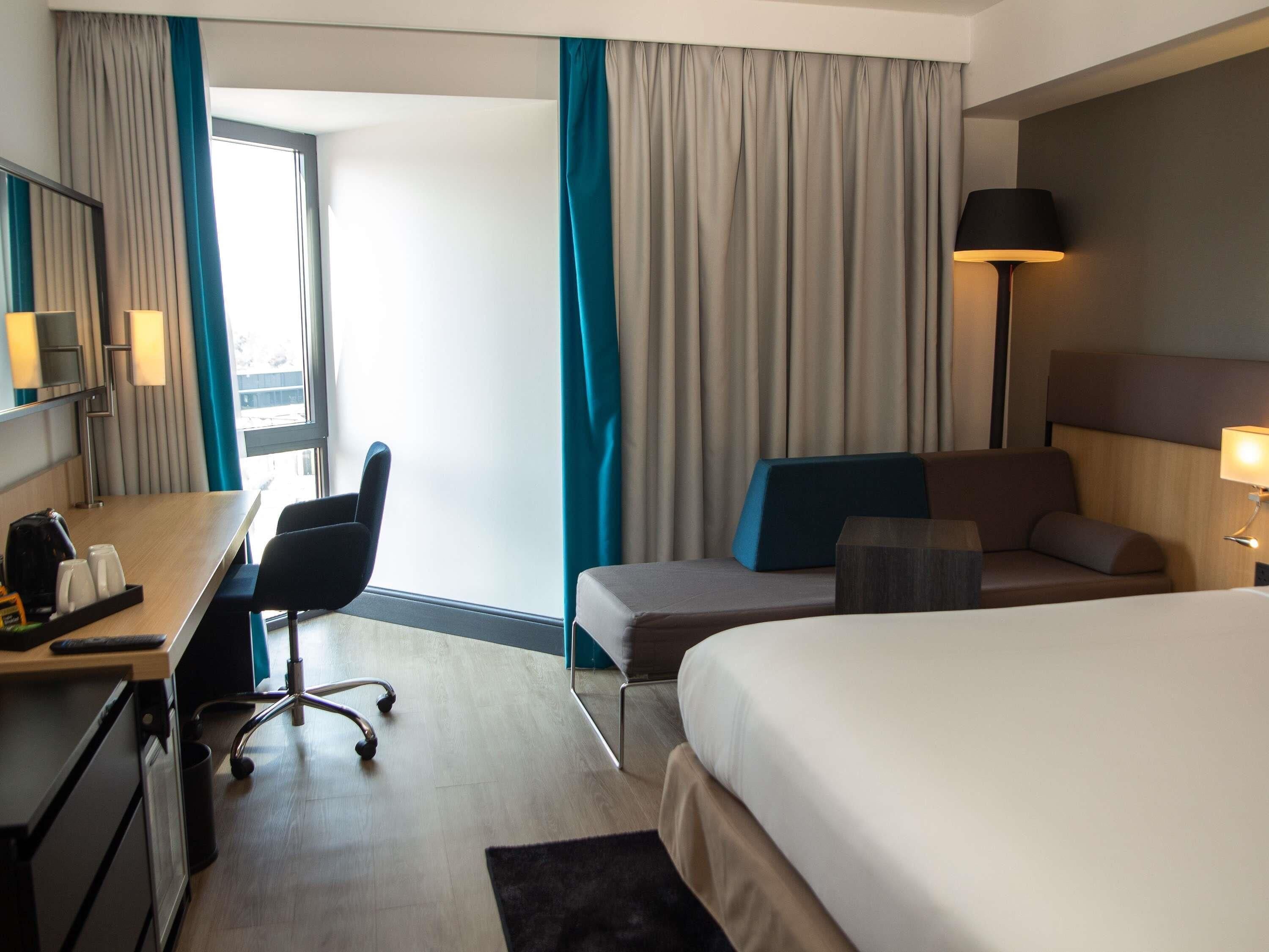 A hotel room with a bed, a desk with a chair, a lamp, and a view of the outdoors.