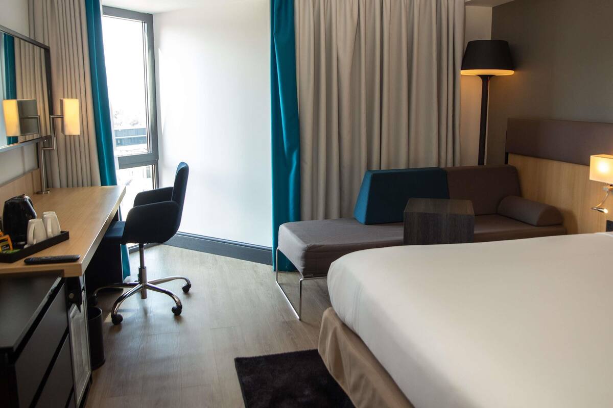 A hotel room with a bed, a desk with a chair, a lamp, and a view of the outdoors.