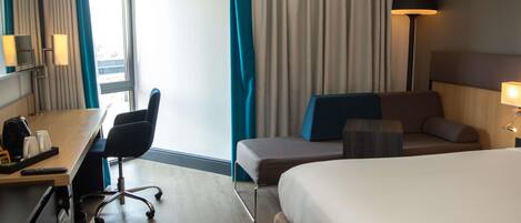 A hotel room with a bed, a desk with a chair, a lamp, and a view of the outdoors.