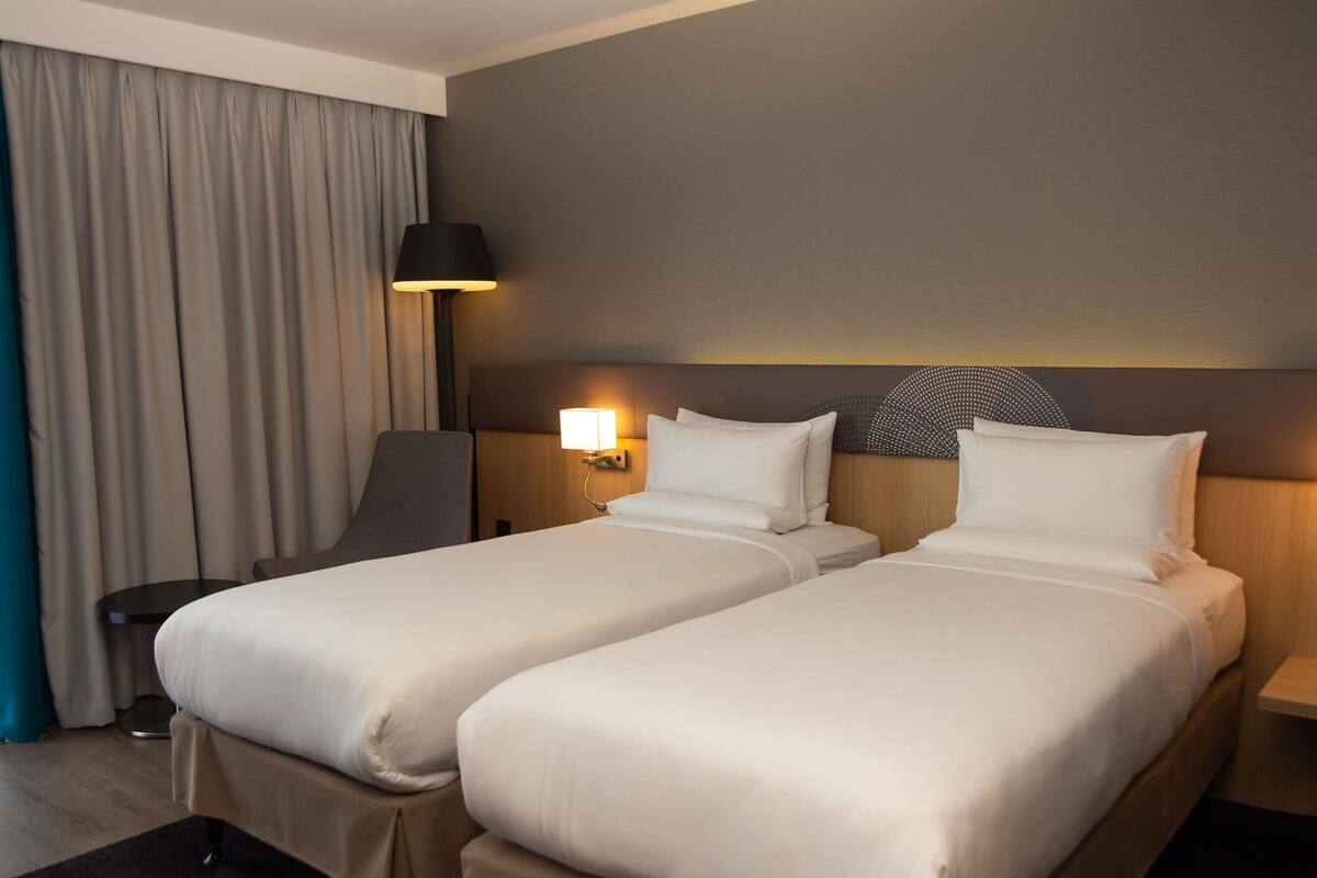 Hotellihuone, jossa on kaksi erillistä sänkyä, työpöytä, tuoli, lamppu ja suuri ikkuna, jossa on verhot.