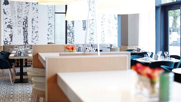 Un restaurant moderne avec un intérieur élégant, caractérisé par de hauts plafonds, de grandes fenêtres et un agencement varié des sièges, comprenant des banquettes et des tables.