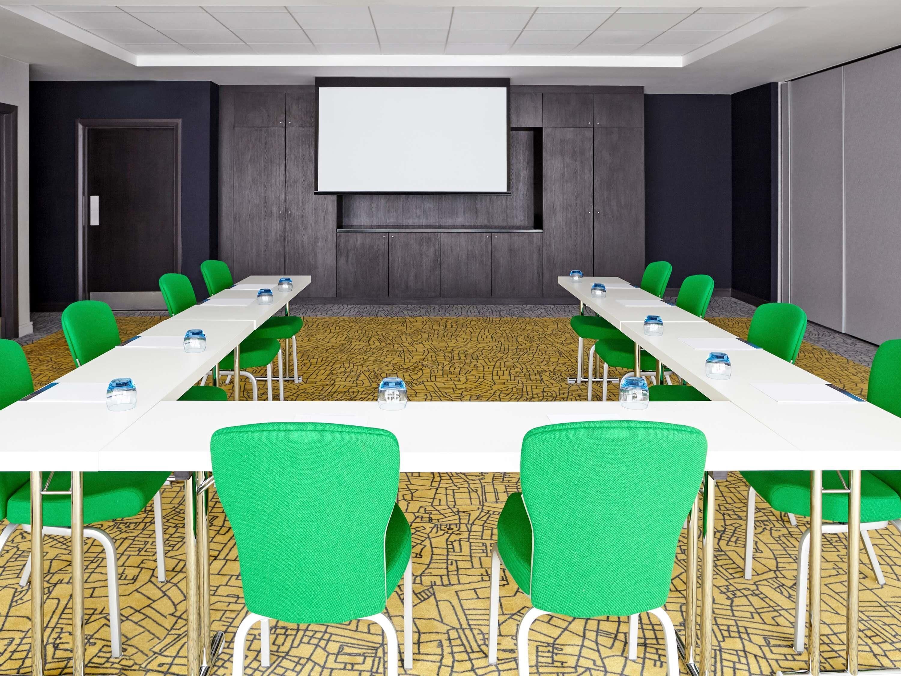Een vergaderzaal met twee lange tafels, groene stoelen, een projectiescherm en een kamerbreed tapijt met een patroon.
