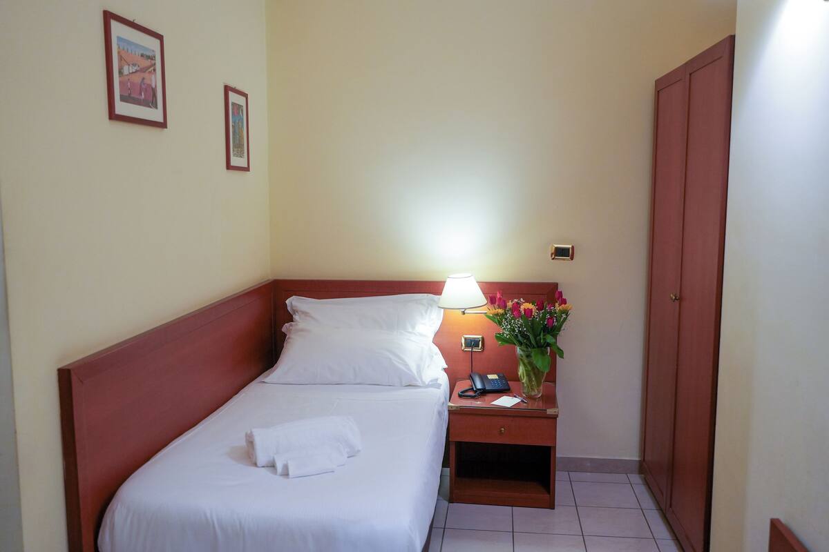 Una camera d'albergo con un letto, un comodino con una lampada, un vaso con fiori e una porta.