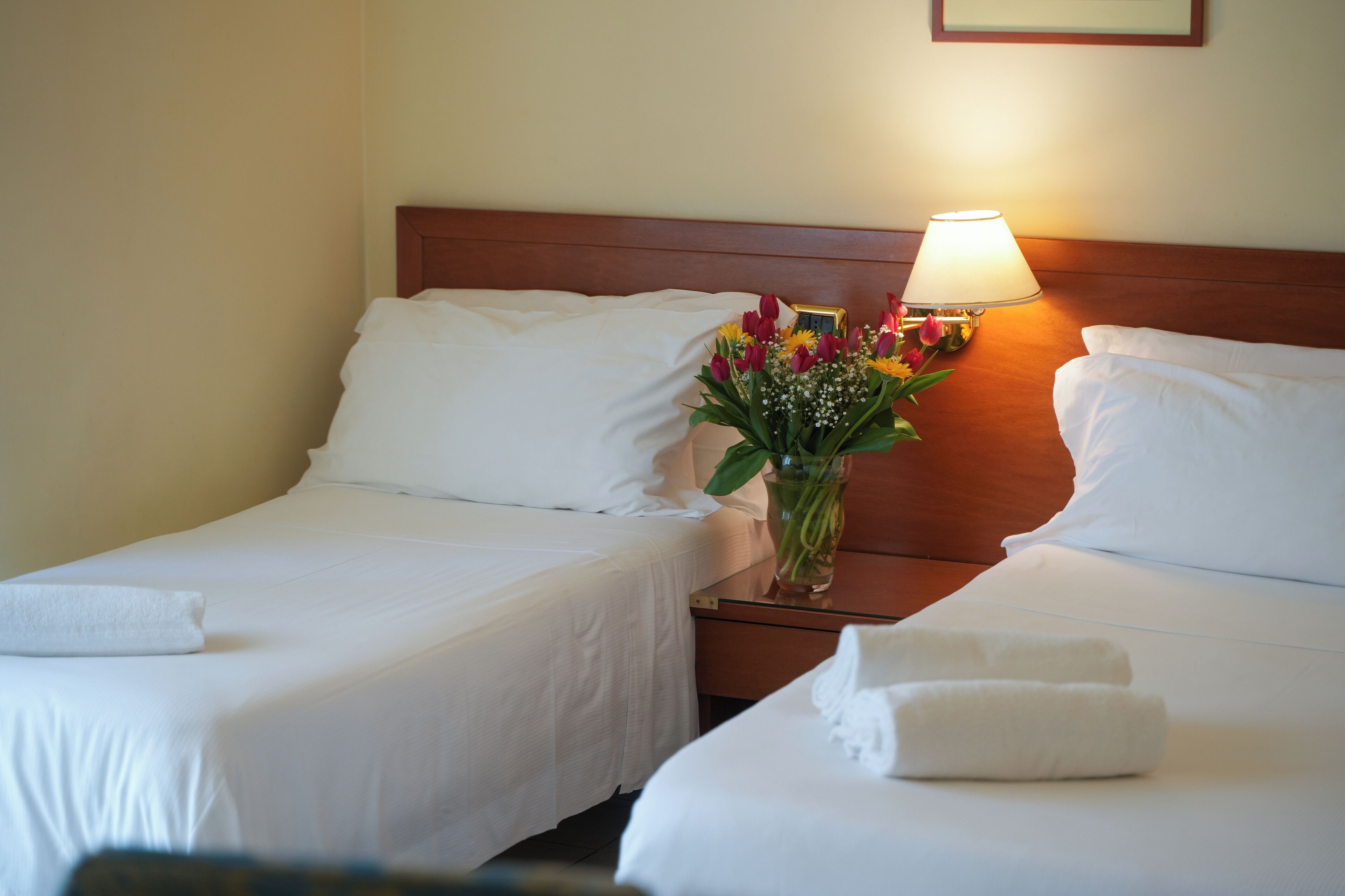 Une chambre d’hôtel avec deux lits simples, du linge de lit blanc, une table de chevet avec une lampe et un vase de fleurs.