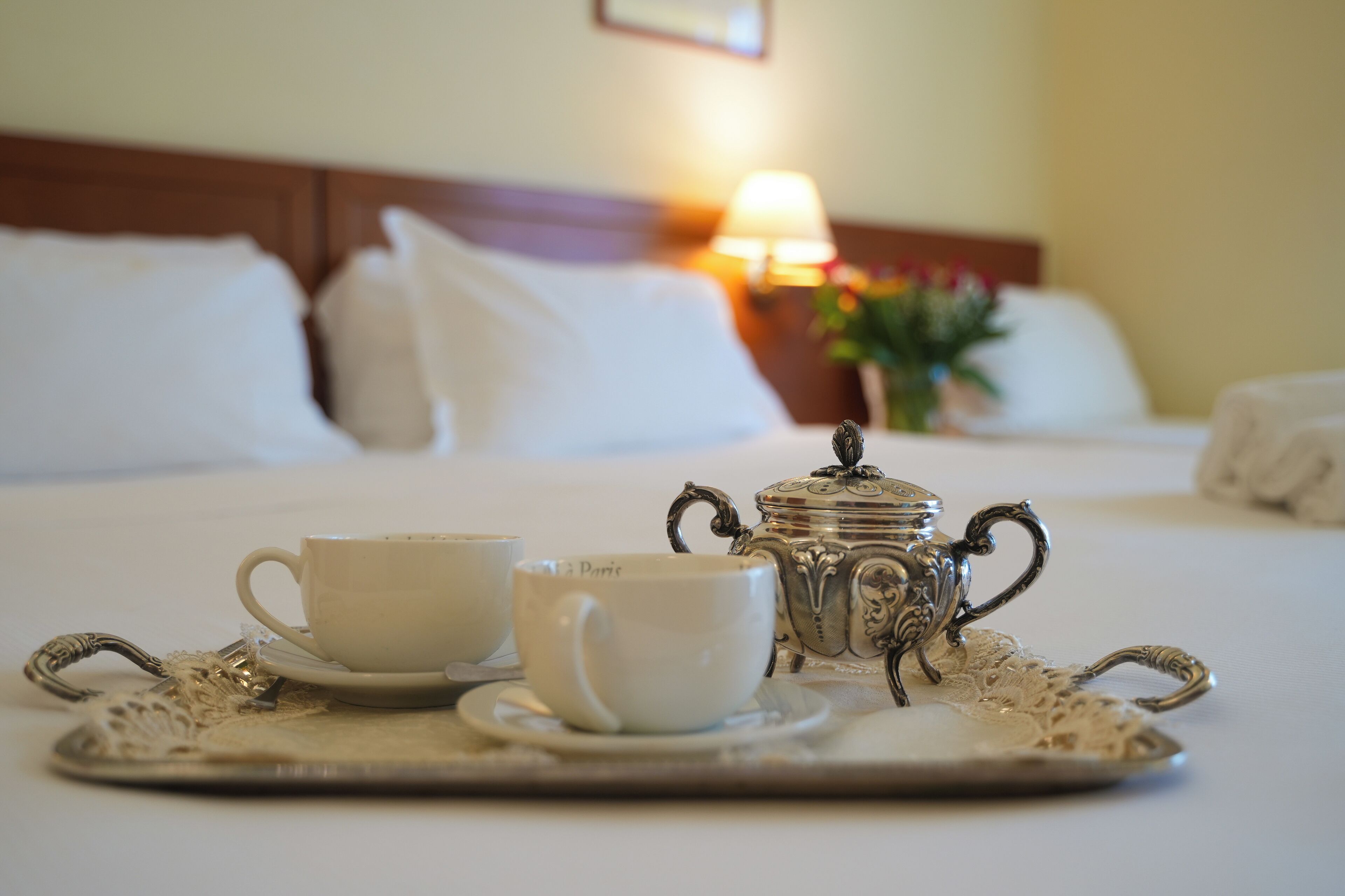 A neatly made bed with white linens and pillows, a bedside table with a lamp, and a tray with a teapot, cups, and a sugar bowl.