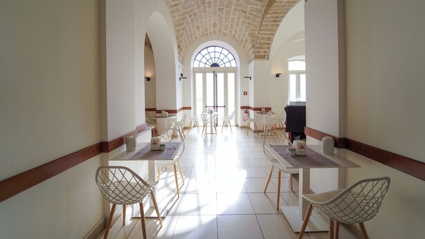 A spacious corridor with high ceilings and arched windows, leading to a dining area with white chairs and tables. - Hotel Adria (Bari)