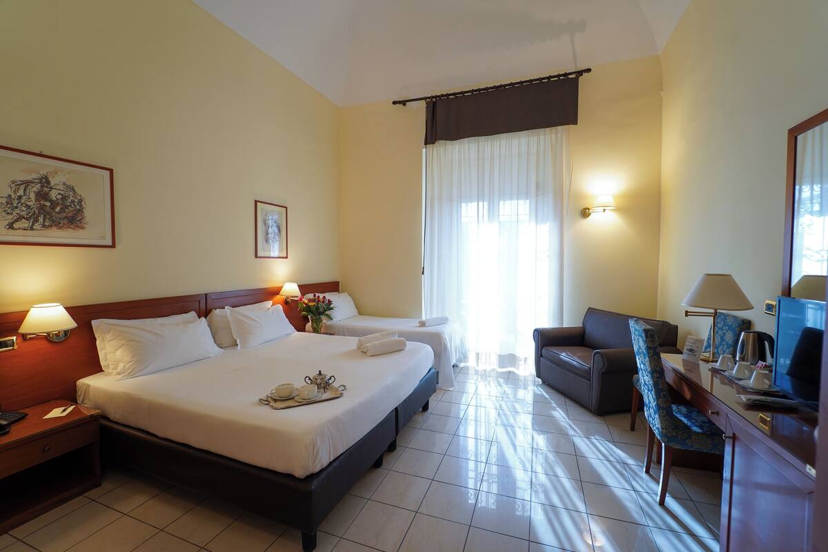 Una camera d'albergo con due letti, una scrivania con un computer, una sedia e un divano.