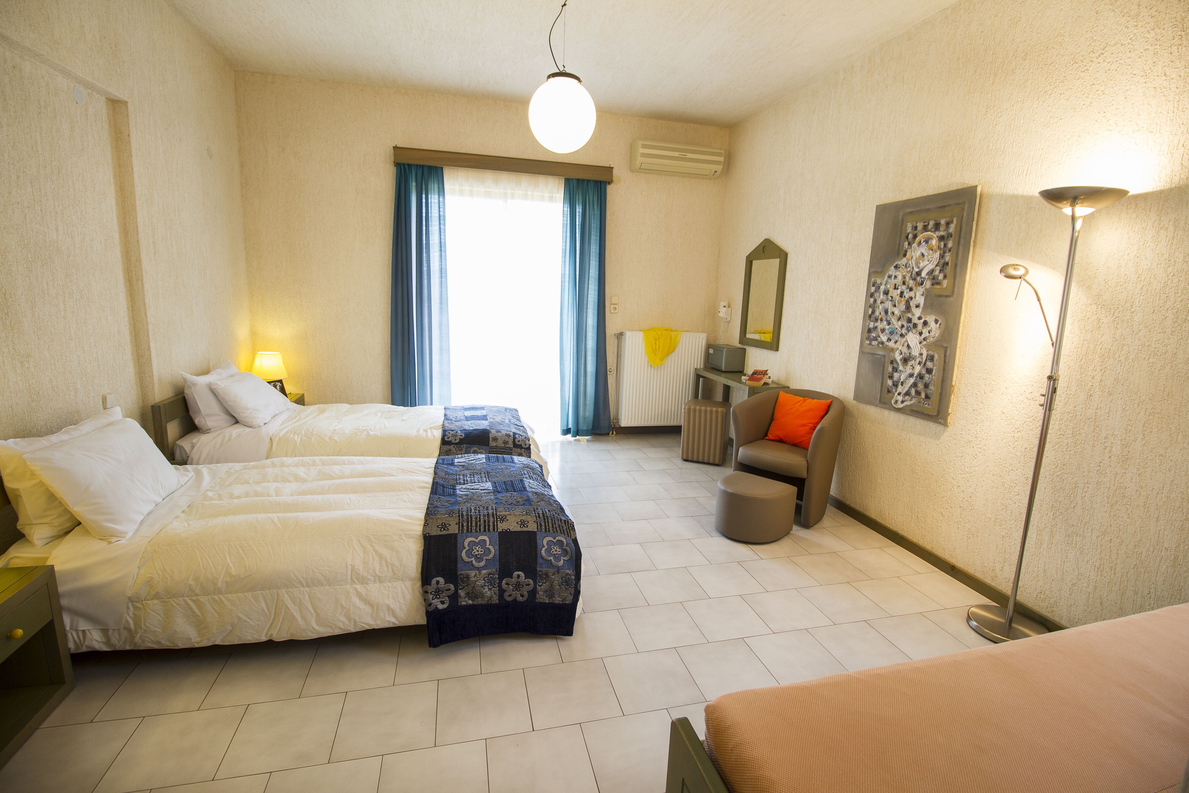 A hotel room with two beds, a desk, a chair, a lamp, and a radiator.
