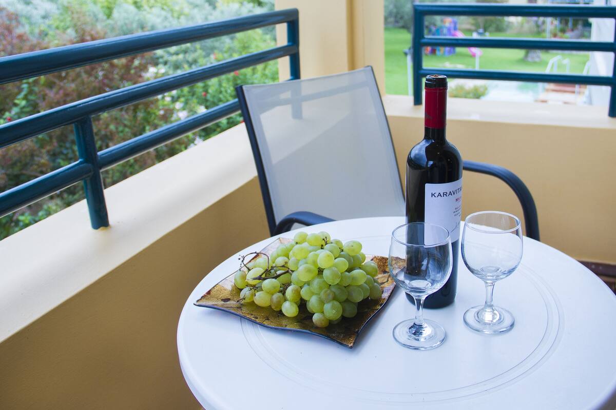Ein Tisch mit einer Karavita Weinflasche, einem Glas und einer Schale grüner Trauben auf einem Balkon mit Blick auf einen Garten.