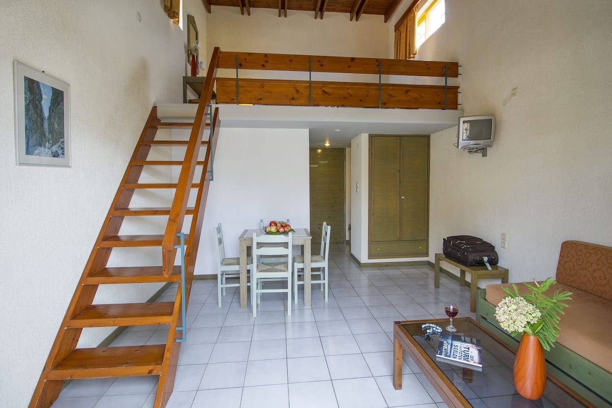 Ein geräumiger Wohnbereich mit einer Holztreppe, einem Esstisch mit Stühlen, einer Couch, einem Couchtisch mit Vase und Zeitschrift sowie einem Fernseher an der Wand.