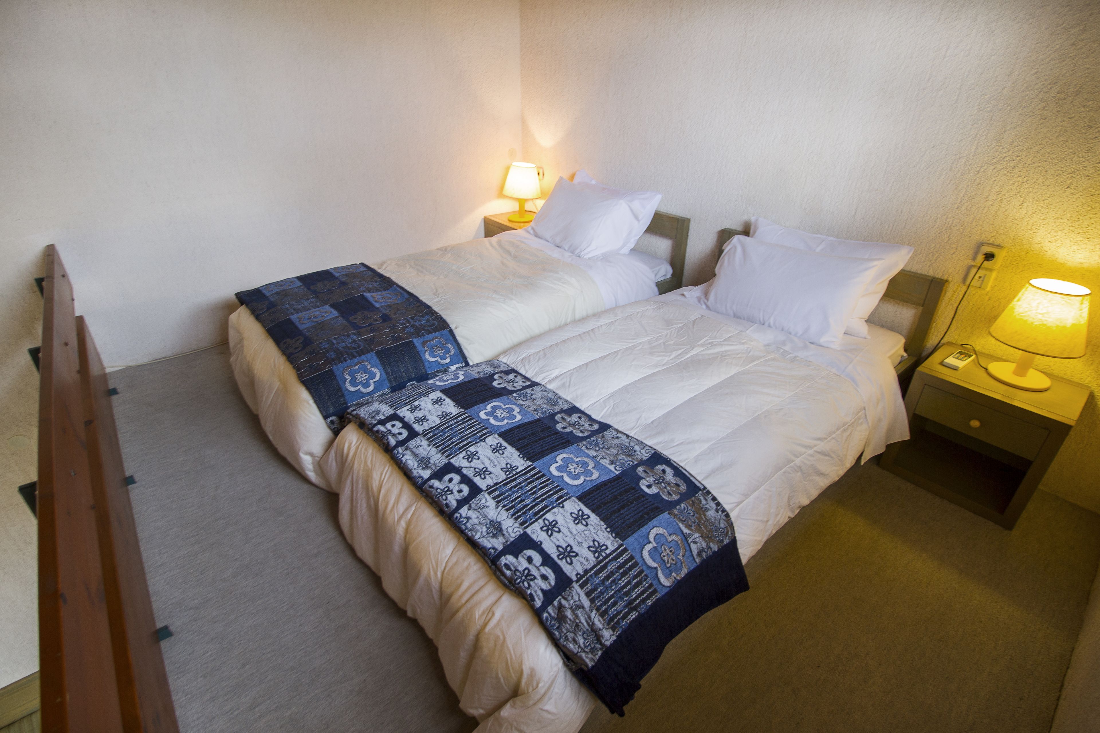 Uma cama de casal com colcha estampada, duas mesas de cabeceira com abajures e uma cadeira de madeira.