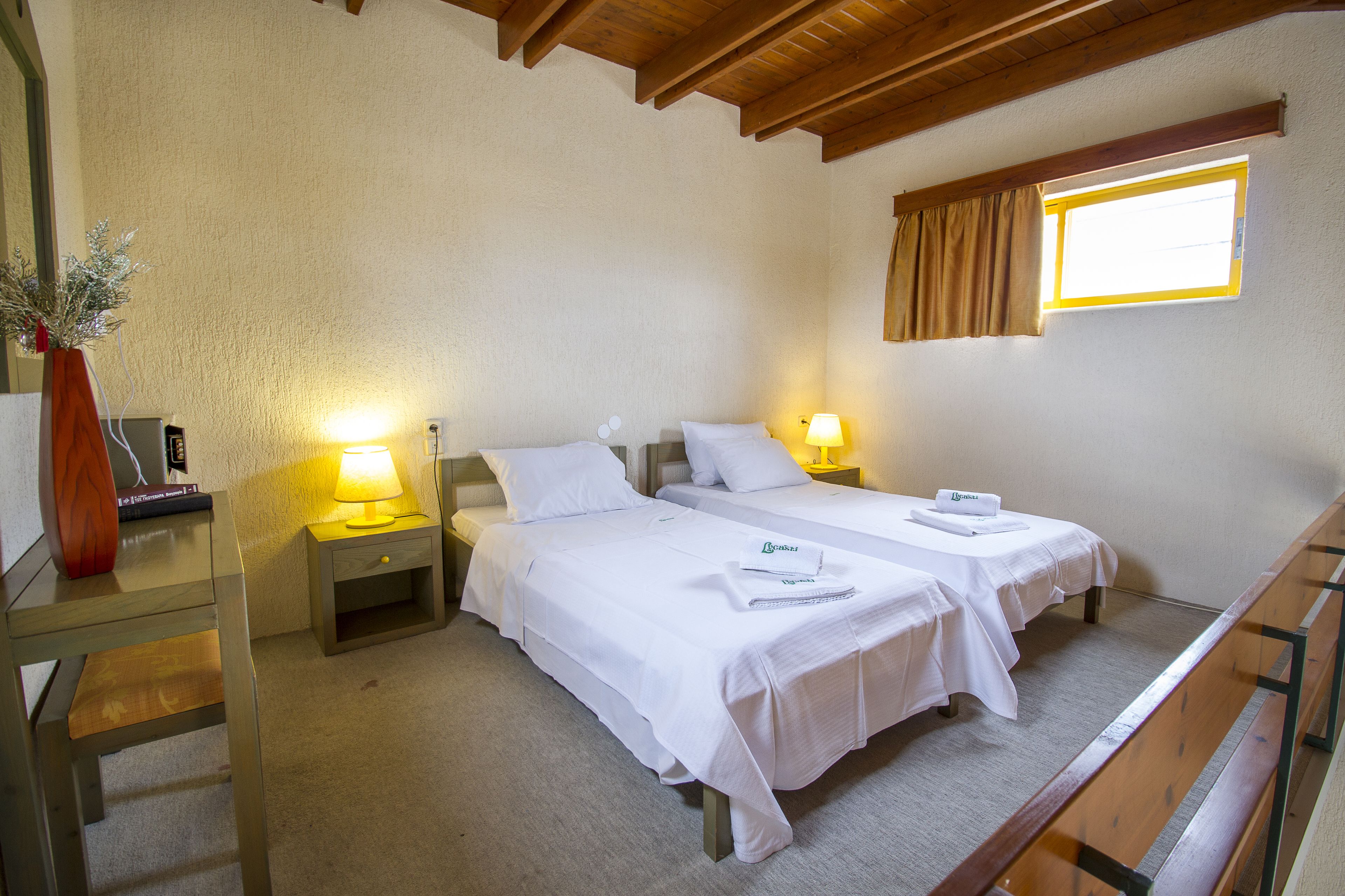 Habitación de hotel con dos camas, un escritorio, una silla, una mesita con lámpara y una ventana con cortinas.