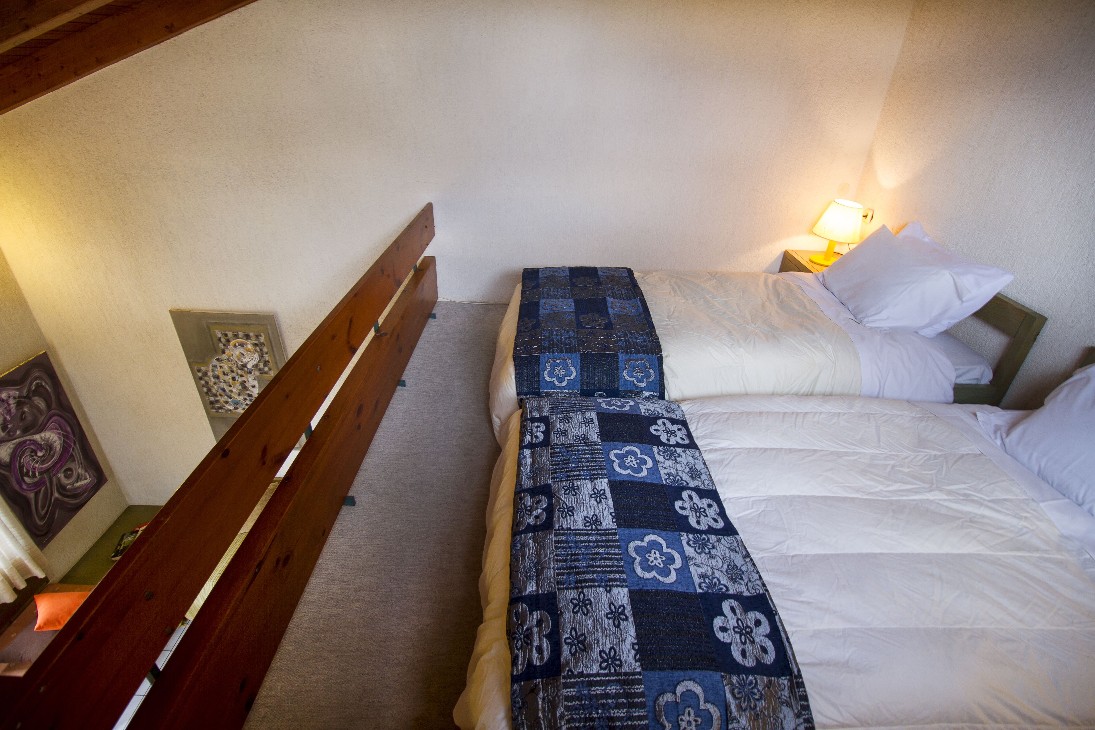 Un dormitorio con dos camas, una con colcha azul y la otra con ropa de cama blanca. Hay un banco de madera y una mesita de noche con una lámpara.
