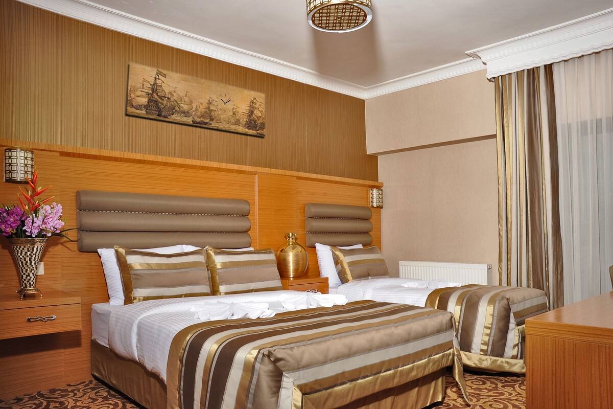 A hotel room with a large bed, two bedside tables, a nightstand, a vase with flowers, a painting on the wall, and a window with curtains.