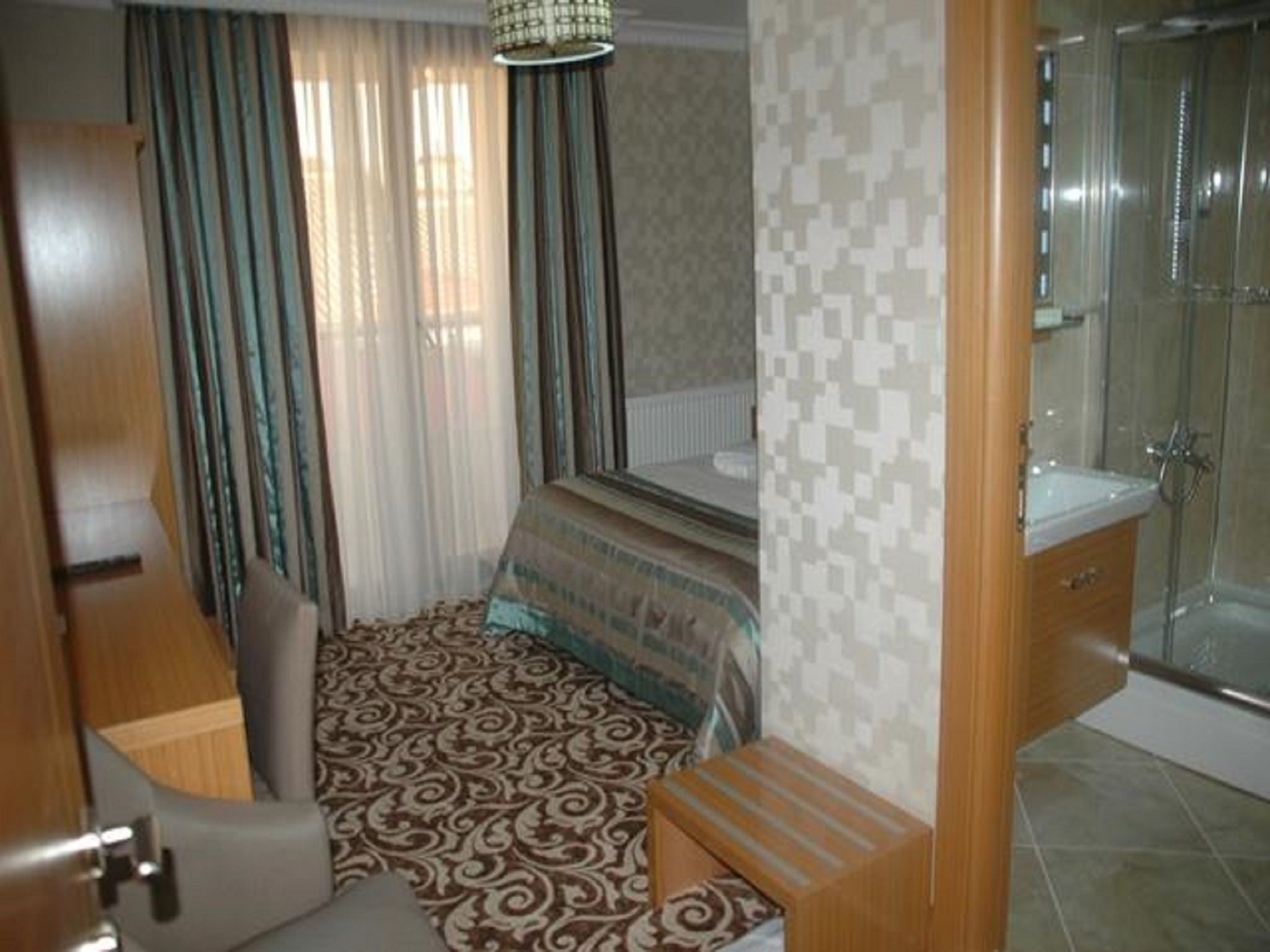Habitación pequeña con cama, una mesita, una silla, zona de ducha y ventana con cortinas.