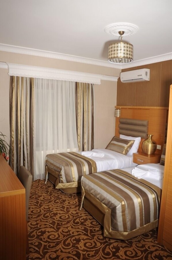 A hotel room with two beds, a desk, a chair, a patterned carpet, a ceiling light fixture, and an air conditioning unit.
