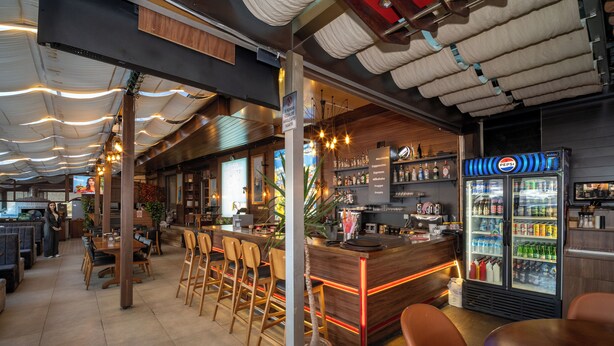Un'area bar moderna con un bancone in legno, sgabelli da bar e una vetrina refrigerata con bevande.
