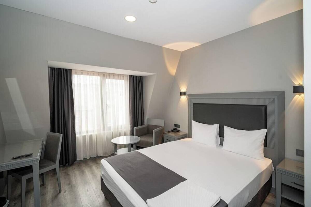 Ein Hotelzimmer mit einem großen Bett, einem Schreibtisch, einem Sessel, einem kleinen Tisch und einem Fenster mit Vorhängen.