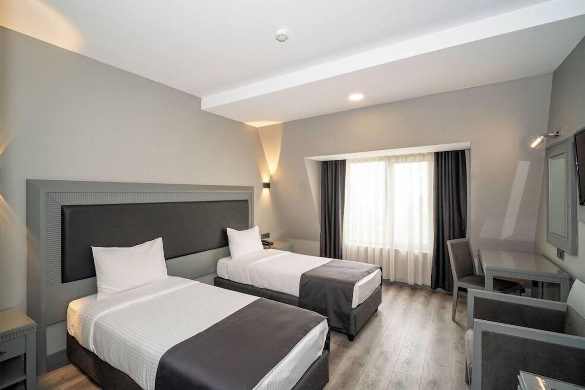 Ein Hotelzimmer mit zwei Betten, einem Schreibtisch, einem Stuhl und einem großen Fenster mit Vorhängen.
