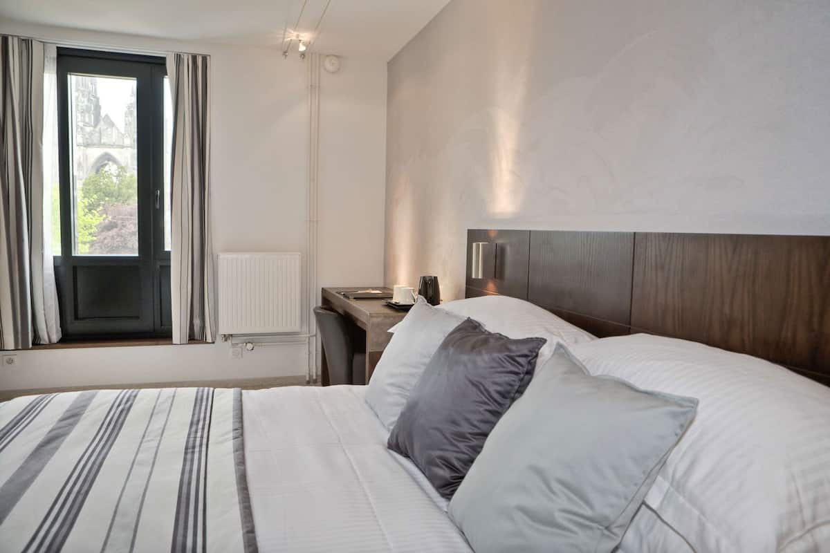 A hotel room with a bed, a desk, a chair, a window with curtains, and a radiator.