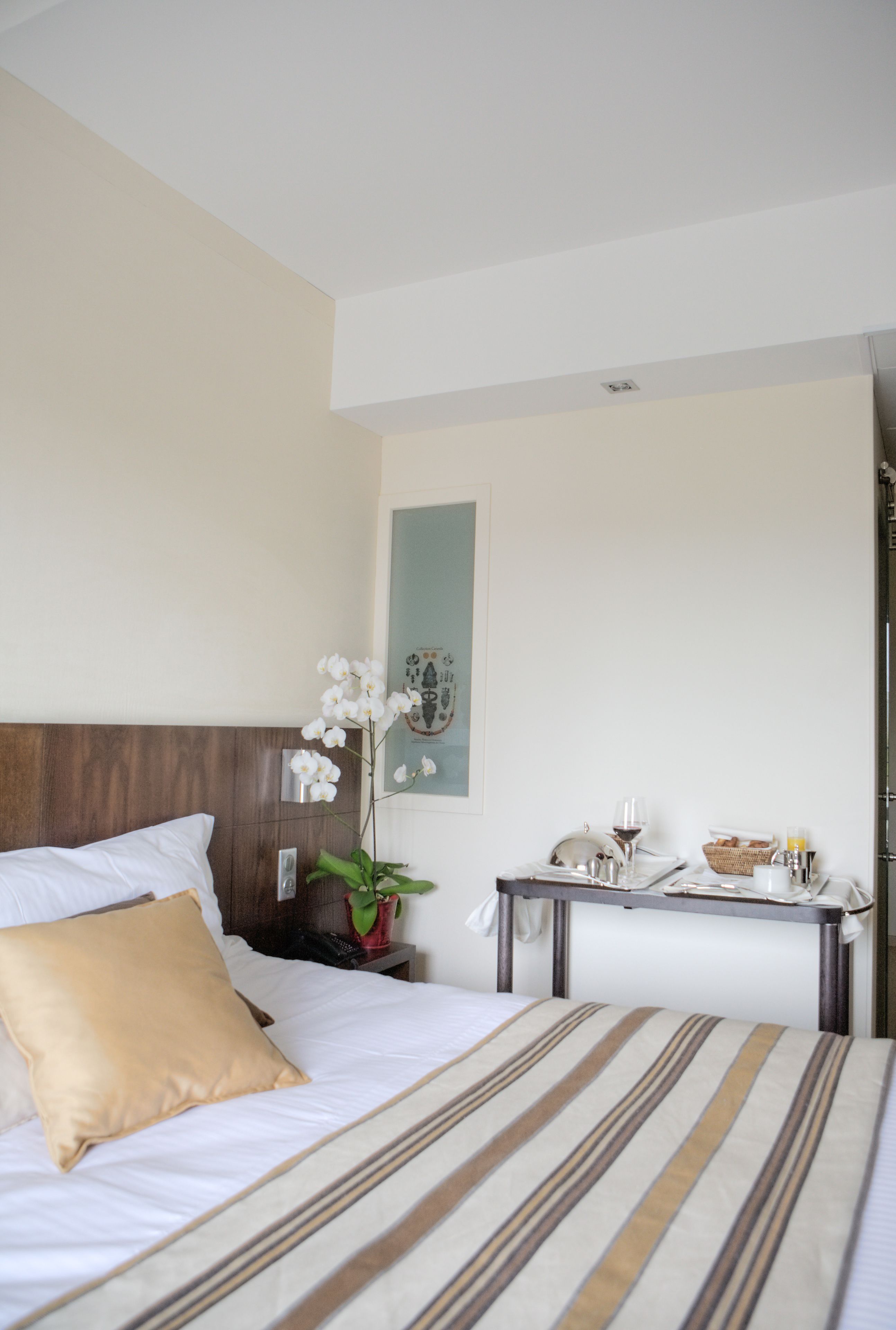 A hotel room with a bed, a bedside table with a vase of flowers, a small table with breakfast items, and a wall-mounted artwork.