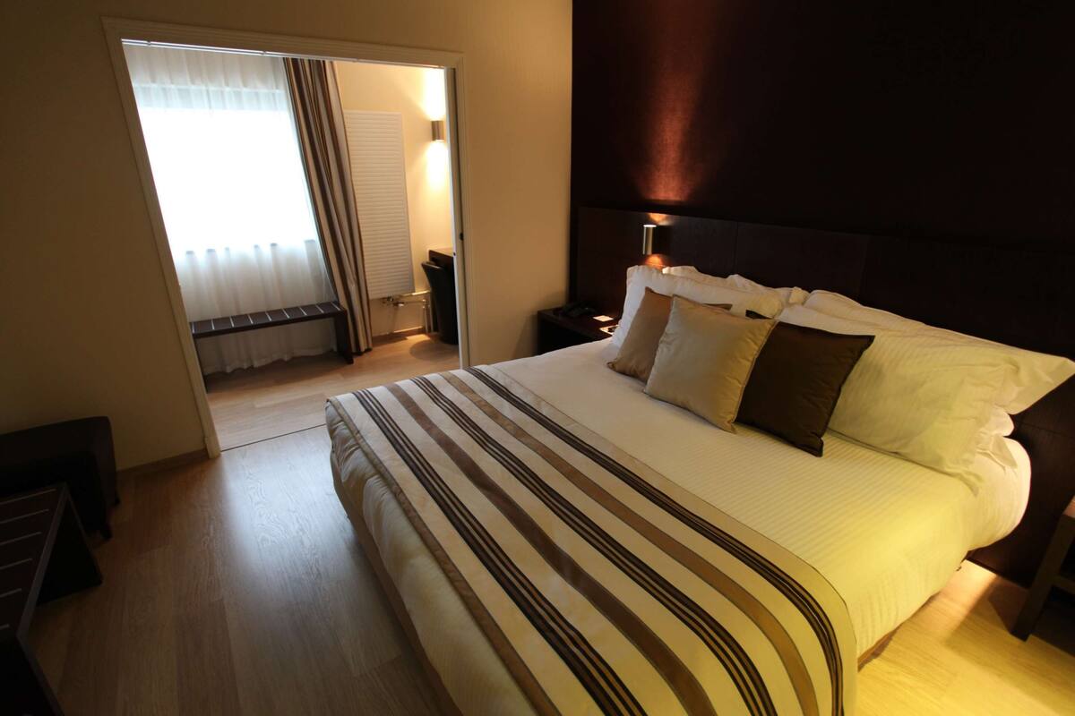 A hotel room with a large bed, a nightstand, a lamp, and a view of a balcony.