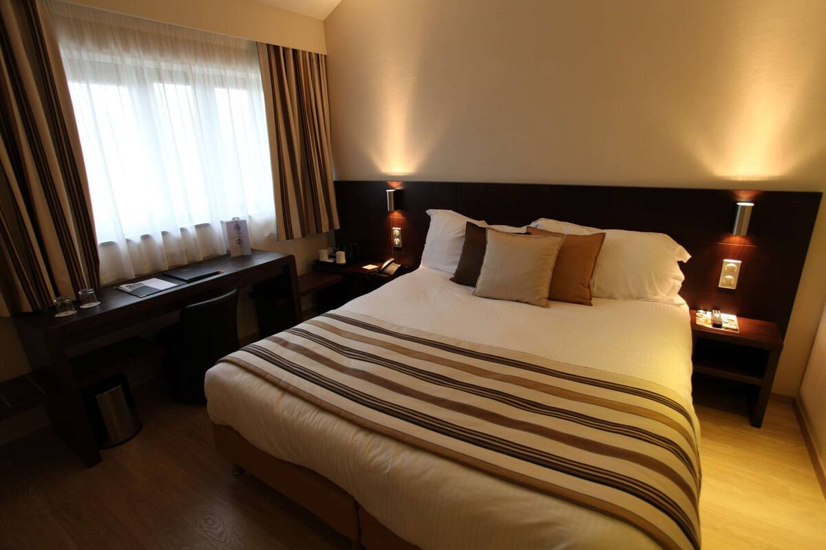 A hotel room with a large bed, a desk, and a chair. There is a nightstand with a lamp and a trash can.
