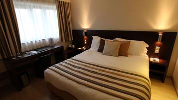 Habitación de hotel con una cama grande, un escritorio y una silla. Se observa una mesita de noche con una lámpara y un bote de basura.