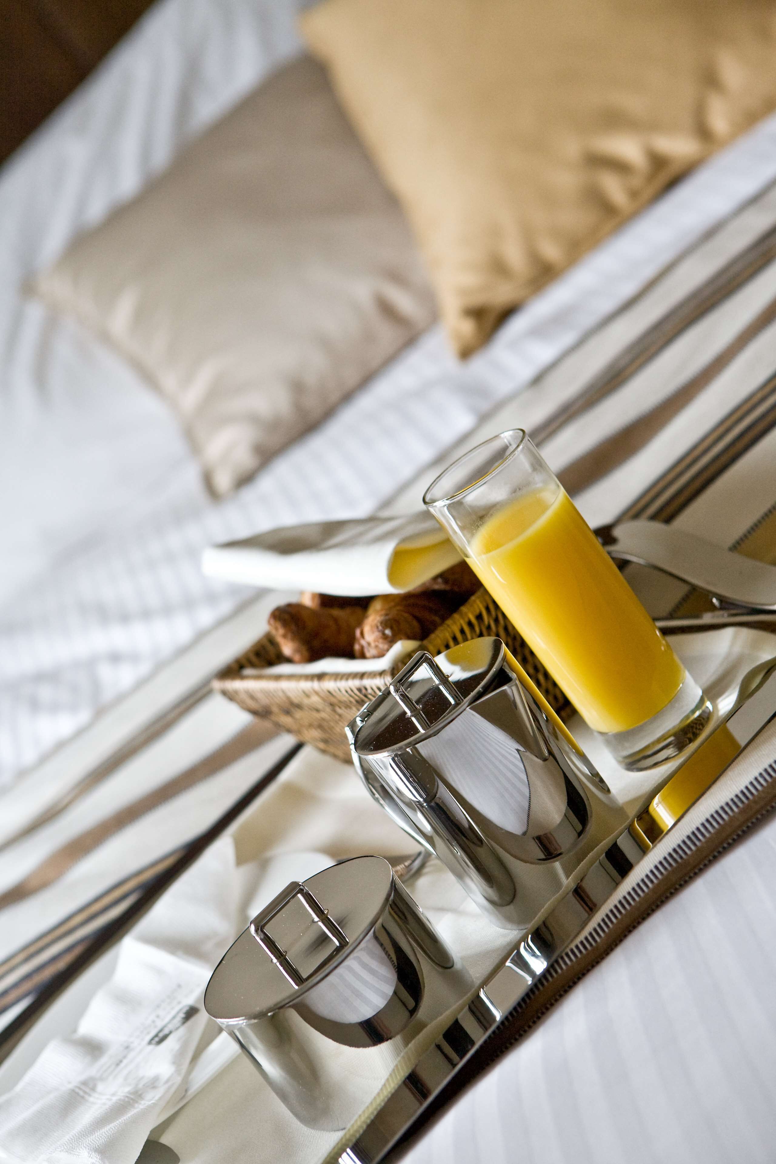 A neatly arranged breakfast tray with a glass of orange juice, a croissant, and silverware on a bed with white linens and a beige pillow.