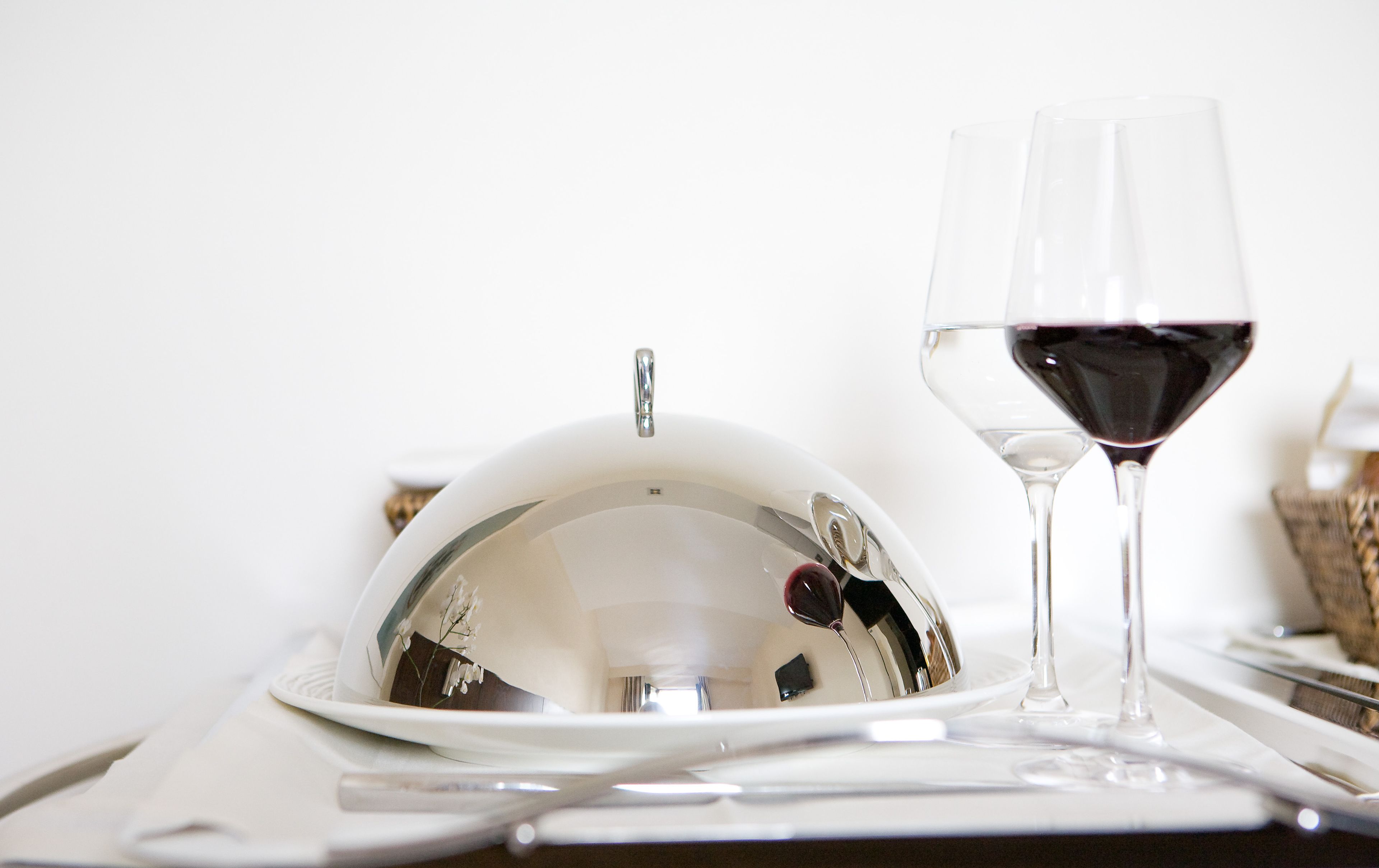 Una mesa de comedor con mantel blanco, una campana de plata, una copa de vino con vino tinto y un tenedor de plata.