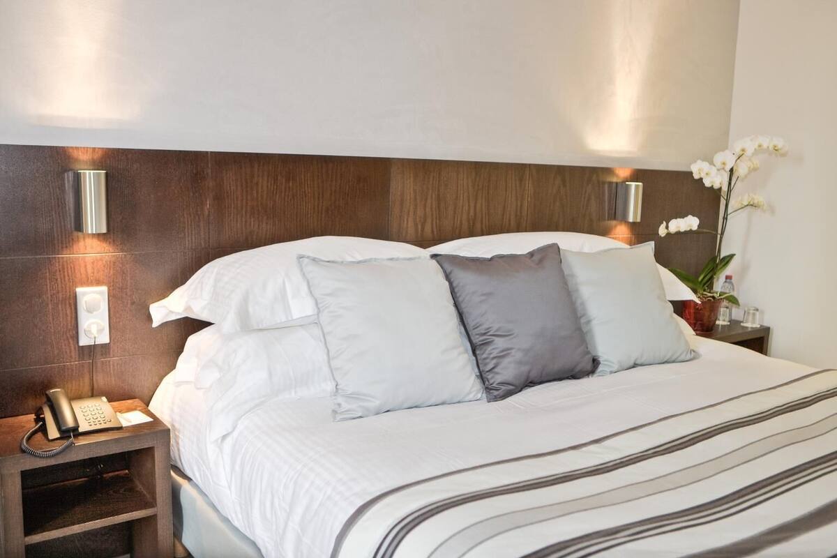 A neatly made bed with white and gray pillows, a bedside phone, and a nightstand with a plant and a water bottle.