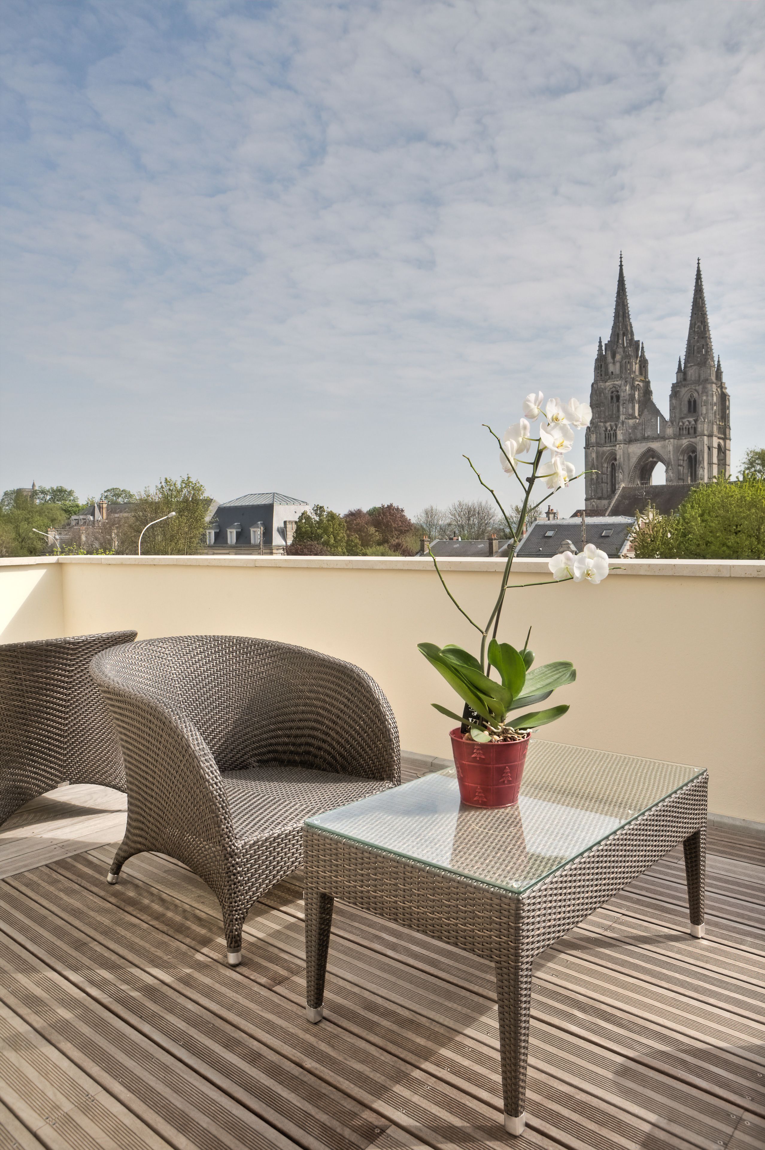 A balcony with wicker furniture, a glass table, and a potted orchid. In the background, there is a church with two spires.