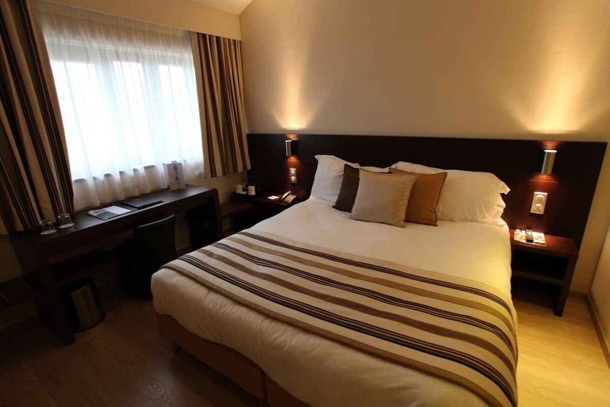 A hotel room with a bed, bedside tables, a desk, a chair, a lamp, and a window with curtains.