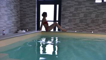 Une personne assise au bord d’une piscine dont les murs sont recouverts de carreaux et qui possède de grandes fenêtres.