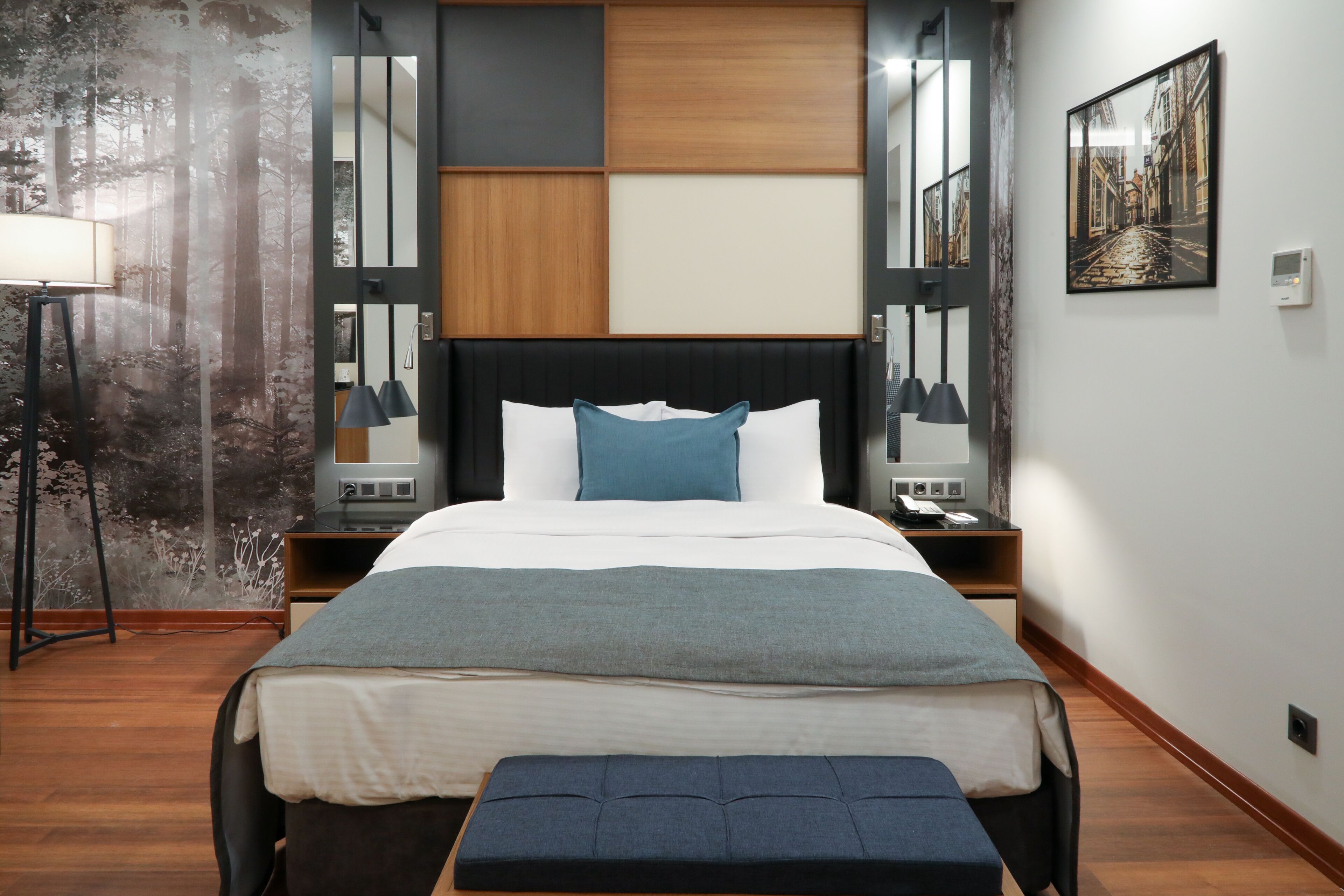 A modern bedroom with a large bed, bedside tables, a bench, a wall-mounted mirror, and a framed picture.