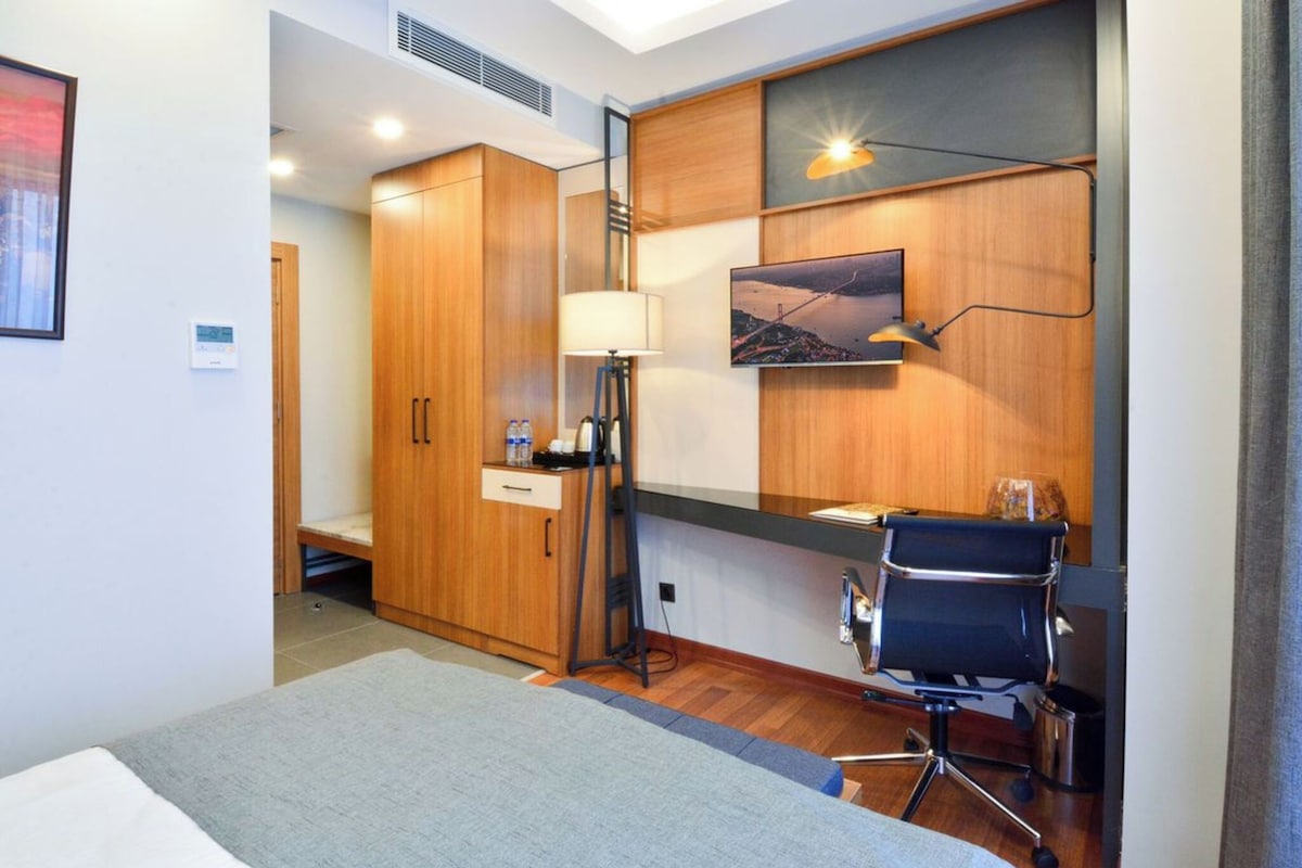 A hotel room with a bed, a desk with a chair, a wardrobe, a lamp, and a painting on the wall.