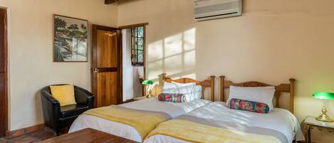 A room with two beds, a wooden table, a chair, a lamp, and a painting on the wall.