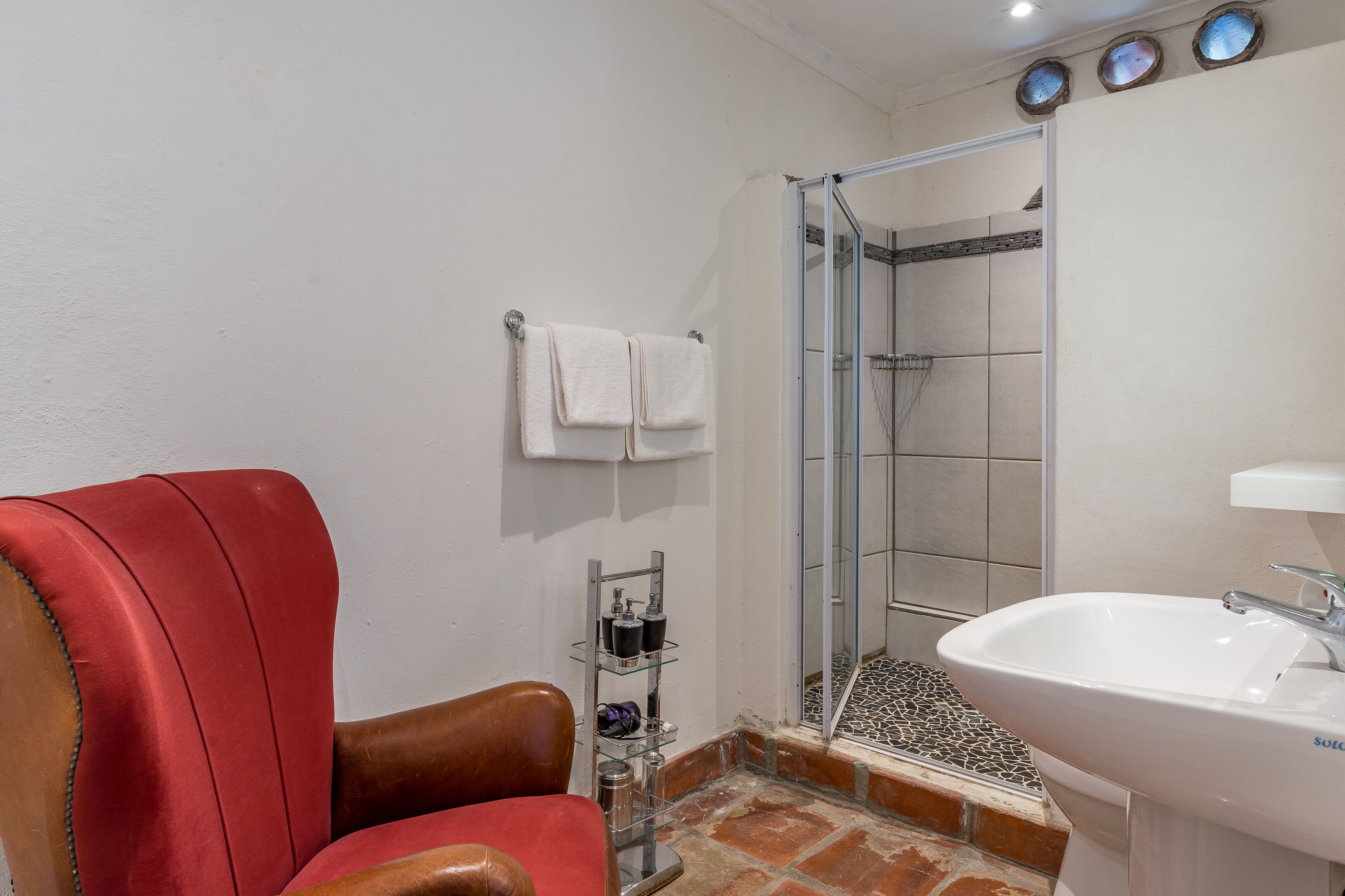A bathroom with a red leather chair, a glass shower enclosure, a white sink, and a tiled shower floor.