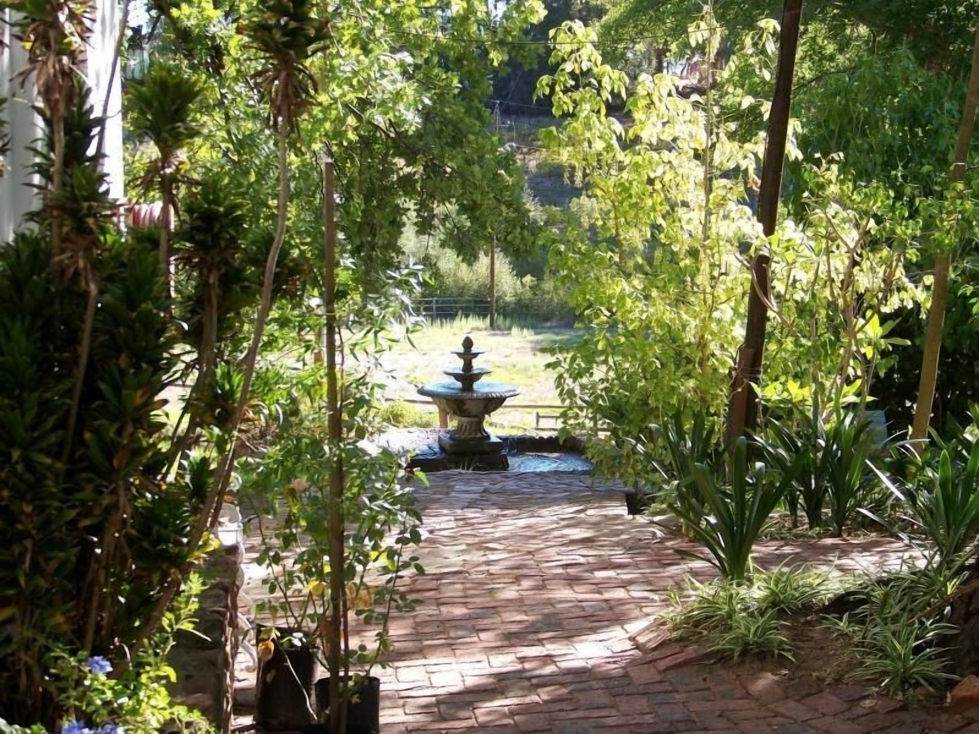 A garden pathway leading to a fountain surrounded by lush greenery and a brick-paved area.