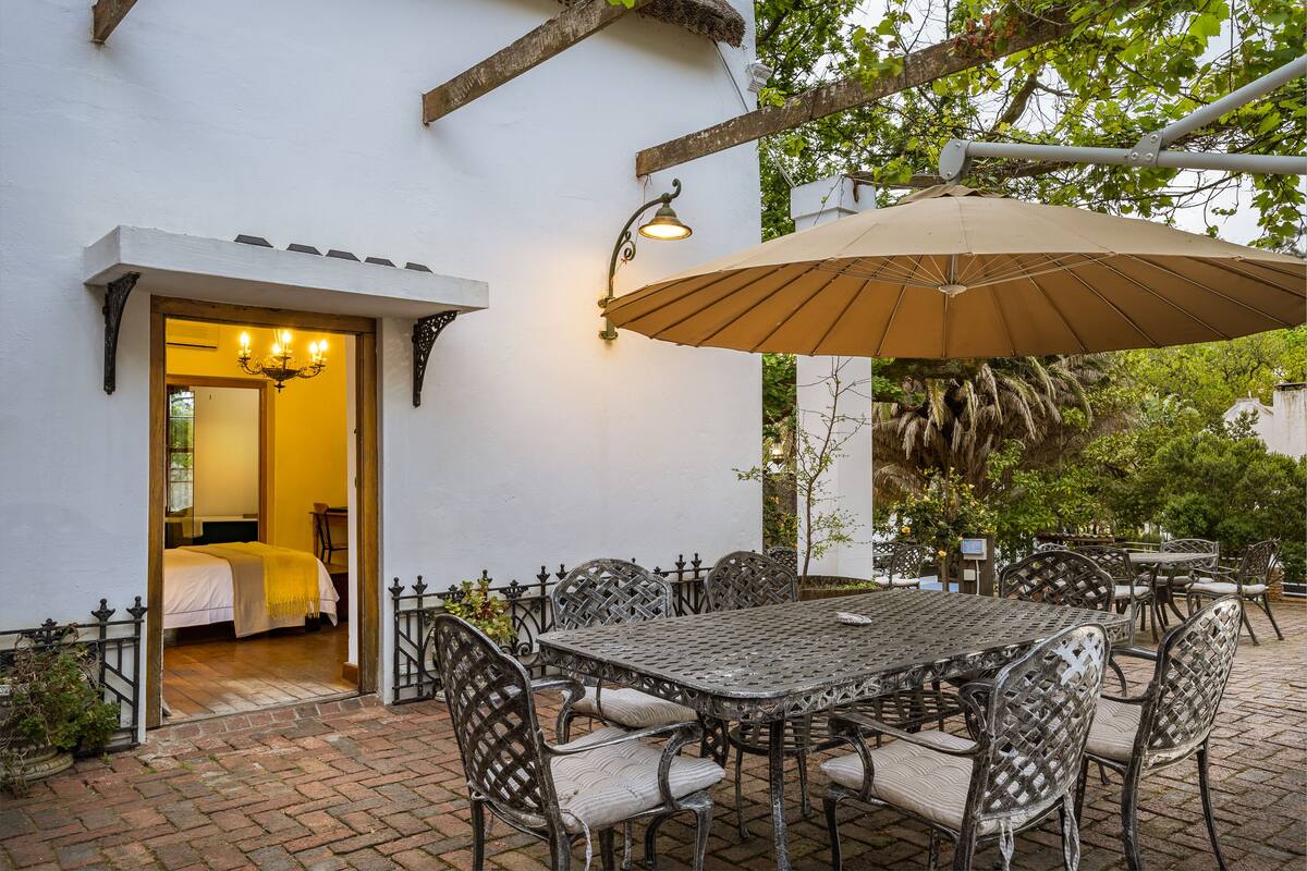 Un patio al aire libre con una mesa y sillas, un gran paraguas y un acceso a una habitación con cama.
