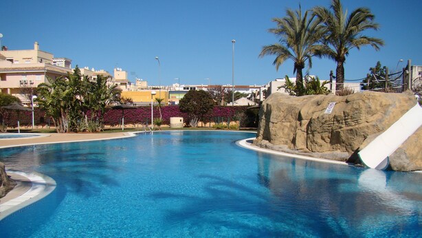 A clear blue pool with a rock feature and palm trees, surrounded by buildings.