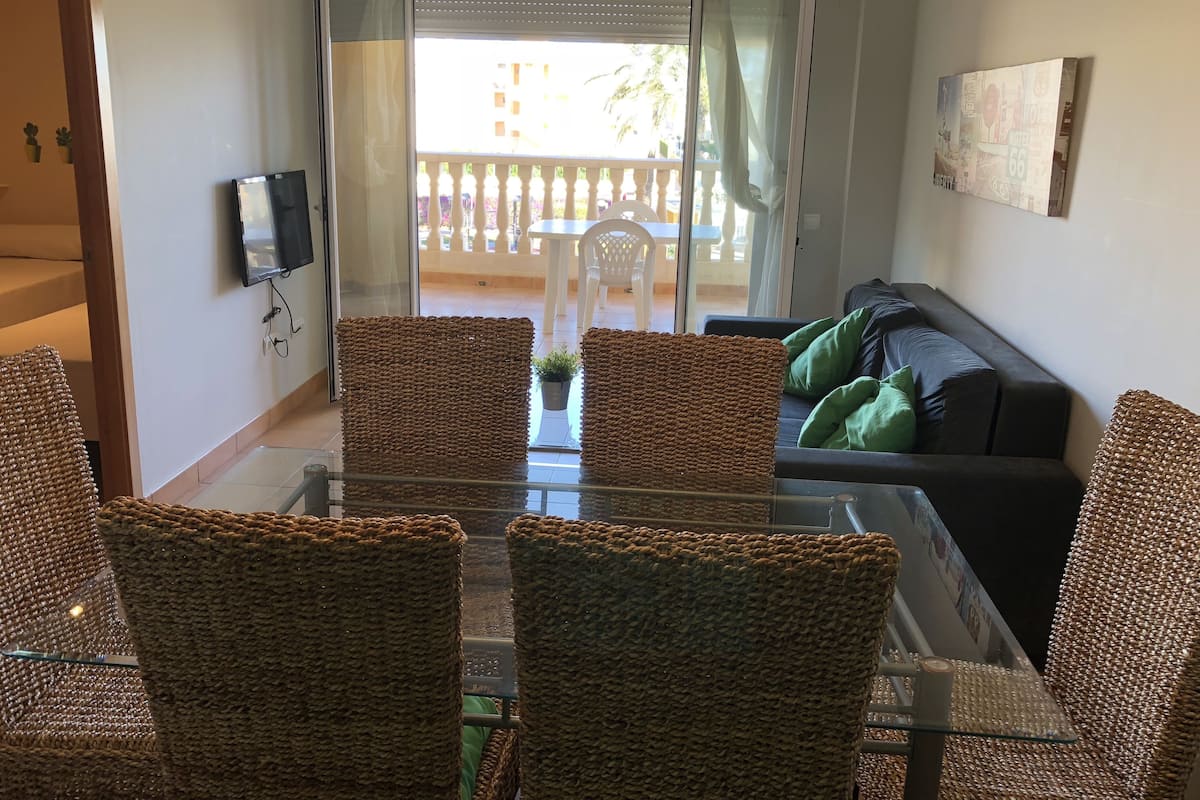 A living area with a glass table and wicker chairs, a black sofa with green cushions, and a balcony with a white railing.