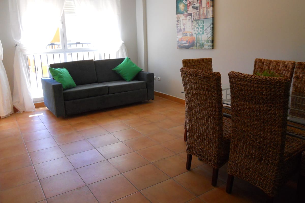 A living room with a sofa, green cushions, a dining table with wicker chairs, and a wall with a collage artwork.