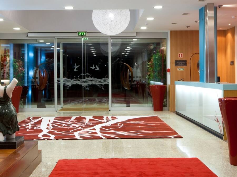 Un vestíbulo de hotel con entrada por puerta de vidrio, alfombra roja, mostrador de recepción y una escultura a la izquierda.