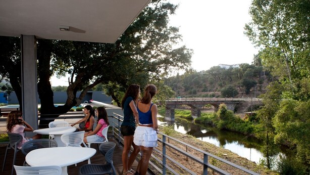 Un patio exterior con mesas y sillas, con vistas a un río y un puente.