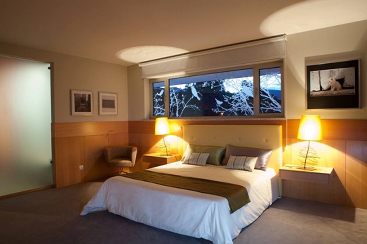 Ein Hotelzimmer mit einem großen Bett, zwei Nachttischlampen, einem Sessel, einem Fenster mit Blick auf Bäume und gerahmten Bildern an der Wand.