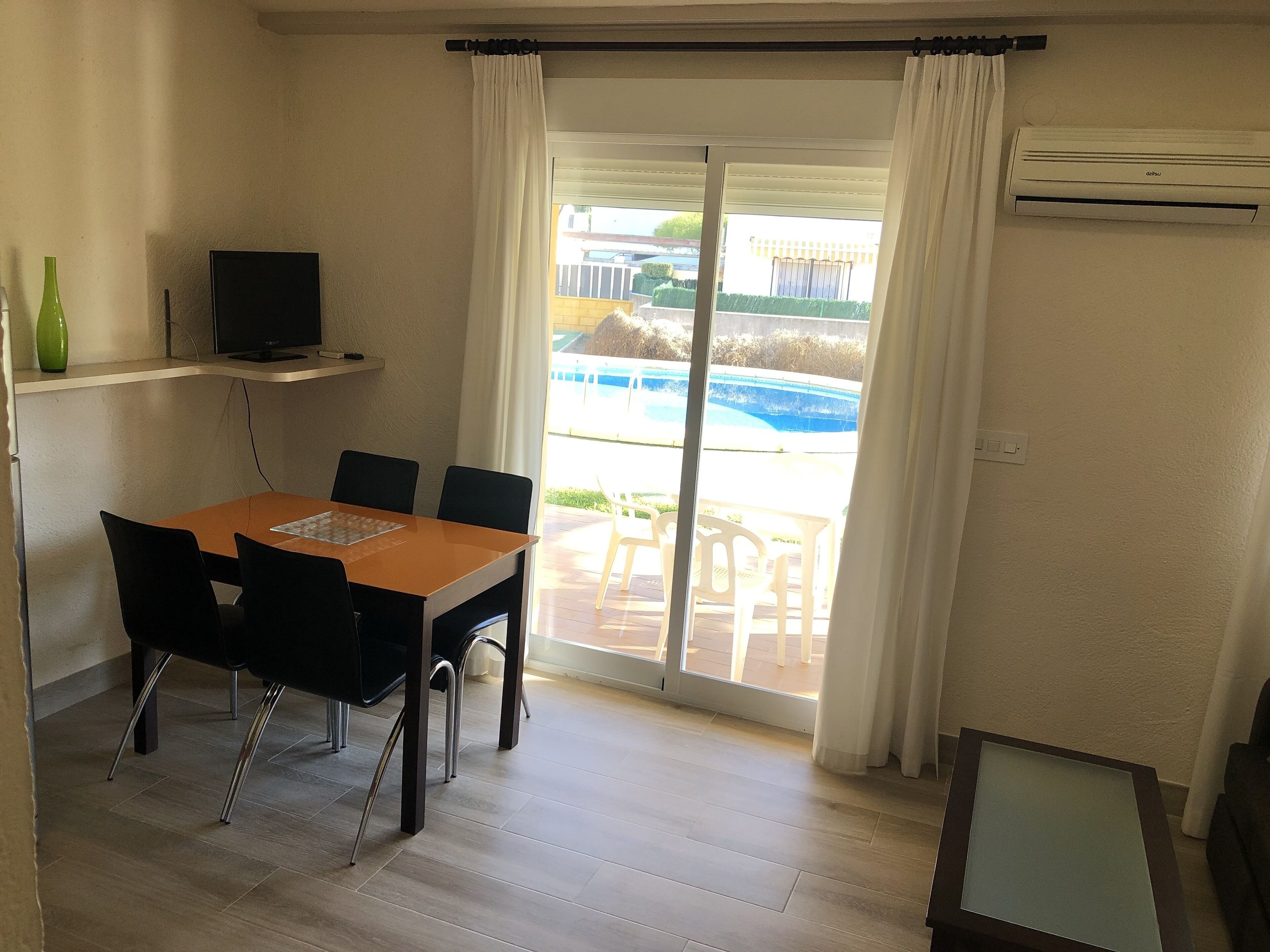 Un coin repas avec une table et des chaises, un téléviseur posé sur une étagère et une vue sur une piscine, visible à travers une porte vitrée.
