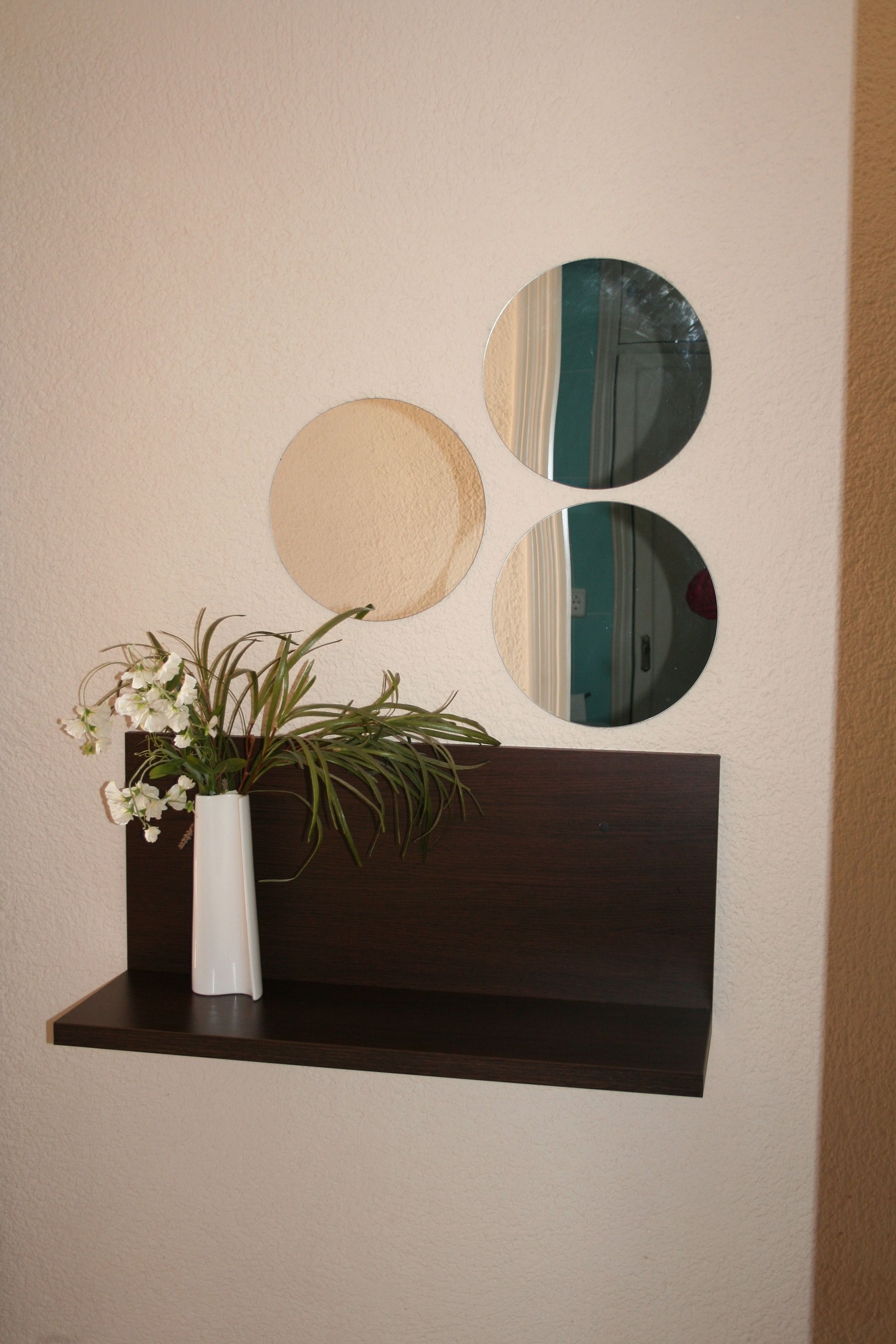 A white vase with flowers on a dark wooden shelf against a wall with three circular mirrors.
