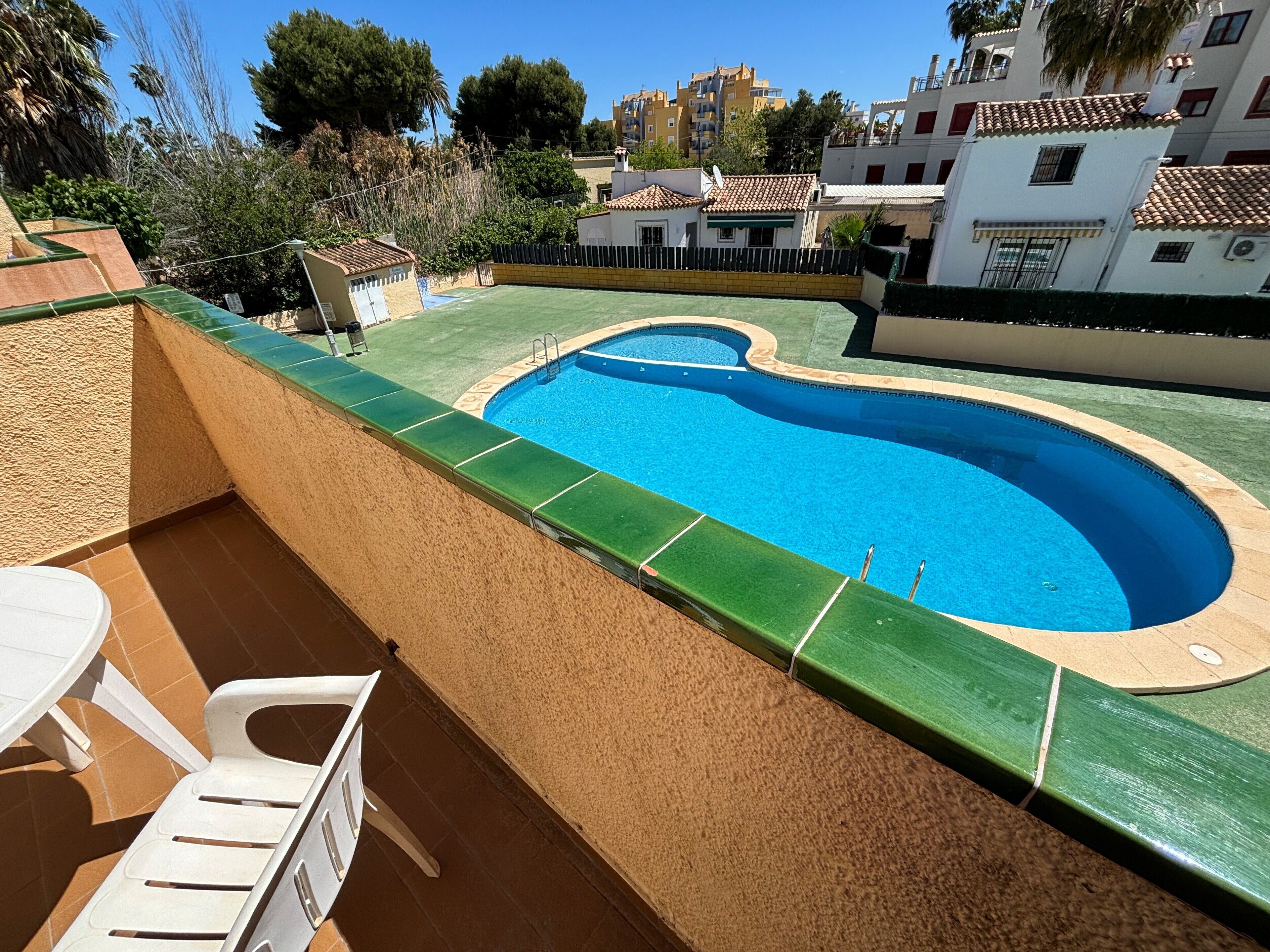 Una piscina con borde verde, rodeada por un muro color beige y un pequeño patio con una silla y una mesa blancos.